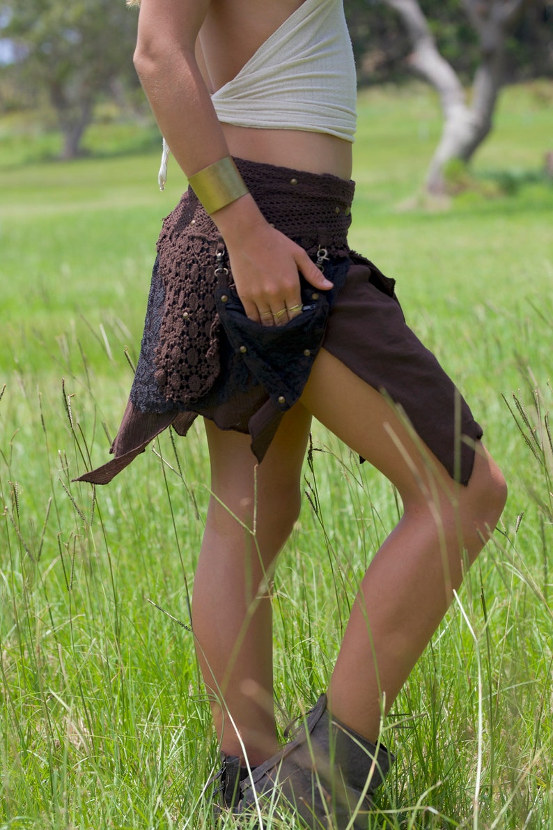 May include: A woman wearing a brown and black skirt with a lace overlay and a gold bracelet. She is standing in a field of tall grass.