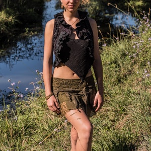 May include: A black sleeveless waistcoat with a ruffled collar and an asymmetrical hem, paired with an olive green mini skirt featuring layered fabric textures, lace detailing, and a distressed finish, worn with black lace-up boots with fringe accents.