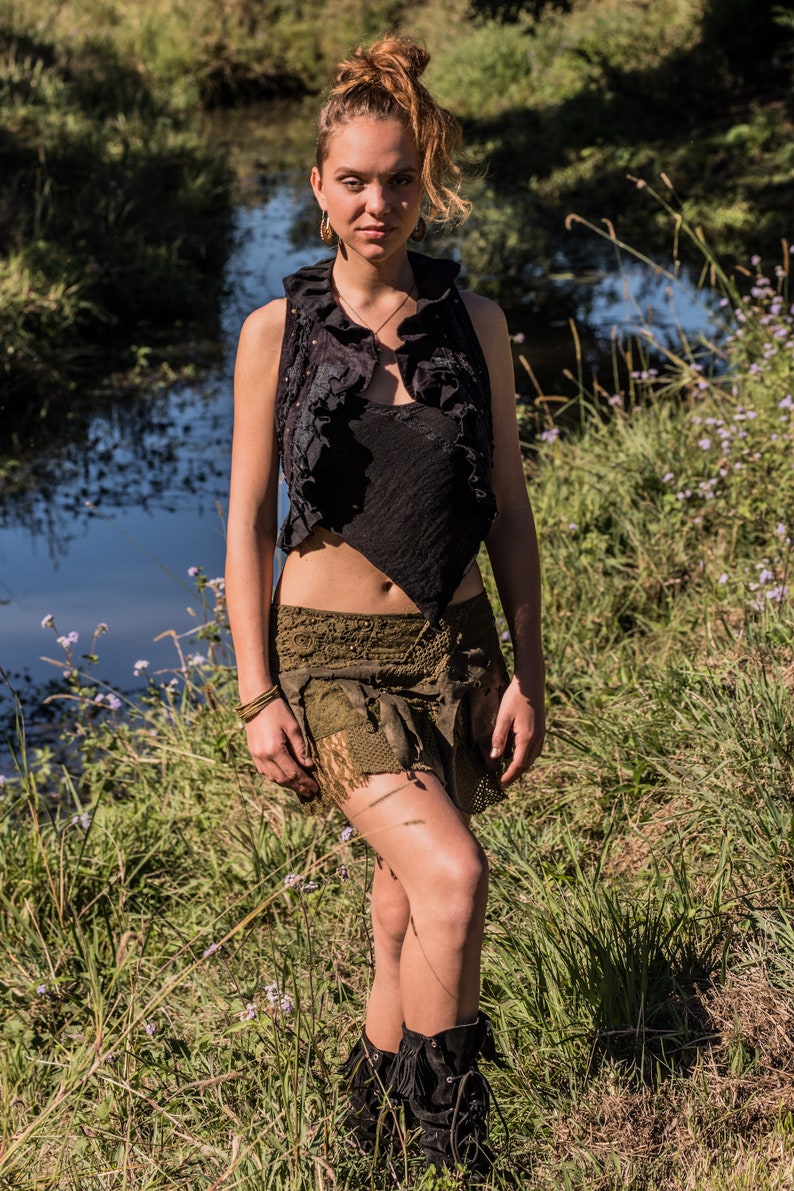 May include: A black sleeveless waistcoat with a ruffled collar and an asymmetrical hem, paired with an olive green mini skirt featuring layered fabric textures, lace detailing, and a distressed finish, worn with black lace-up boots with fringe accents.