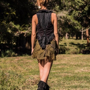 May include: A black sleeveless vest with a ruffled collar and a lace-up back closure, paired with an olive green layered mini skirt featuring mesh and lace textures, and black mid-calf boots with fringe detailing on the upper portion.