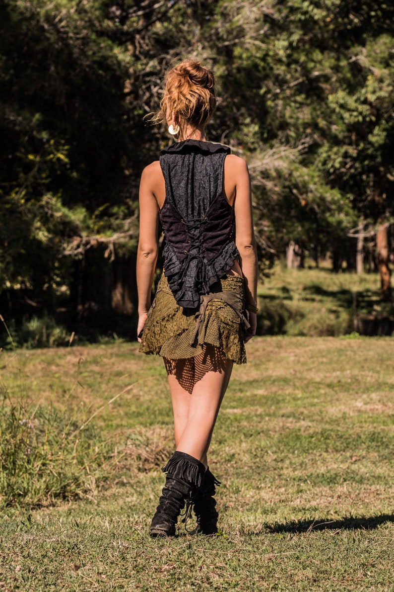May include: A black sleeveless vest with a ruffled collar and a lace-up back closure, paired with an olive green layered mini skirt featuring mesh and lace textures, and black mid-calf boots with fringe detailing on the upper portion.