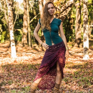 May include: A woman wearing a teal top with a lace-up front and a burgundy lace skirt. She is standing in a forest with fall leaves on the ground.