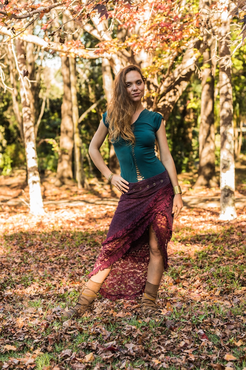 May include: A woman wearing a teal top with a lace-up front and a burgundy lace skirt. She is standing in a forest with fall leaves on the ground.