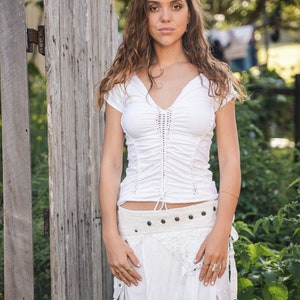 May include: A woman wearing a white crochet top with a white crochet skirt and a brown leather belt. She is standing in front of a wooden fence.
