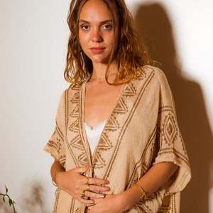 Puede incluir: Una mujer lleva un poncho de punto beige con un patr&oacute;n geom&eacute;trico. El poncho tiene cuello en V y est&aacute; abierto por delante. La mujer lleva anillos de oro en los dedos y una pulsera de oro en la mu&ntilde;eca.