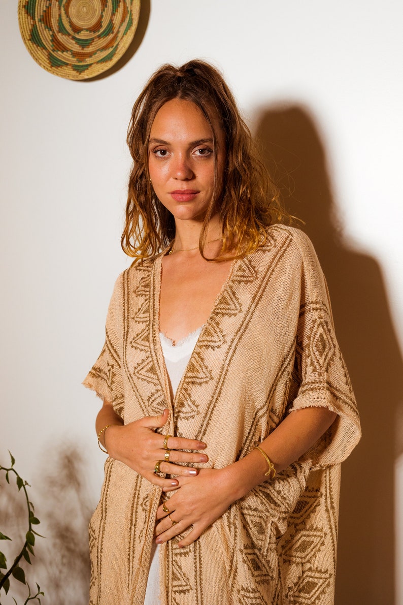 Puede incluir: Una mujer lleva un poncho de punto beige con un patr&oacute;n geom&eacute;trico. El poncho tiene cuello en V y est&aacute; abierto por delante. La mujer lleva anillos de oro en los dedos y una pulsera de oro en la mu&ntilde;eca.