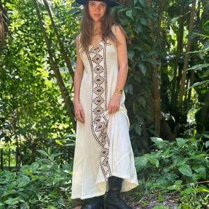 May include: A white maxi dress with brown geometric embroidery. The dress has a V-neckline and a sleeveless design. The model is wearing a black hat and black boots.