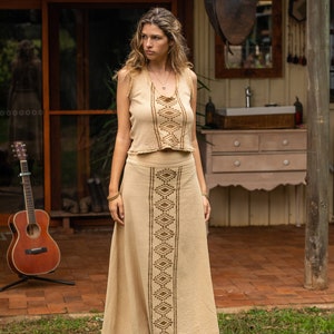 May include: A woman wearing a beige sleeveless top and matching skirt with brown geometric embroidery. The skirt is long and flowing, and the top is cropped. She is standing in front of a wooden building with a saddle hanging from the ceiling.
