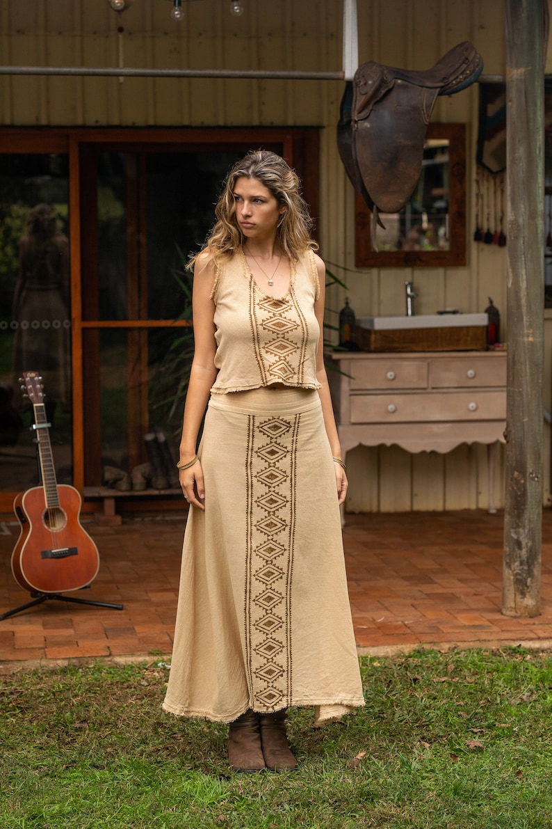 May include: A woman wearing a beige sleeveless top and matching skirt with brown geometric embroidery. The skirt is long and flowing, and the top is cropped. She is standing in front of a wooden building with a saddle hanging from the ceiling.