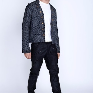 Puede incluir: Un hombre con una chaqueta estampada en negro y gris con botones dorados, pantalones vaqueros negros con costuras decorativas y zapatos negros.