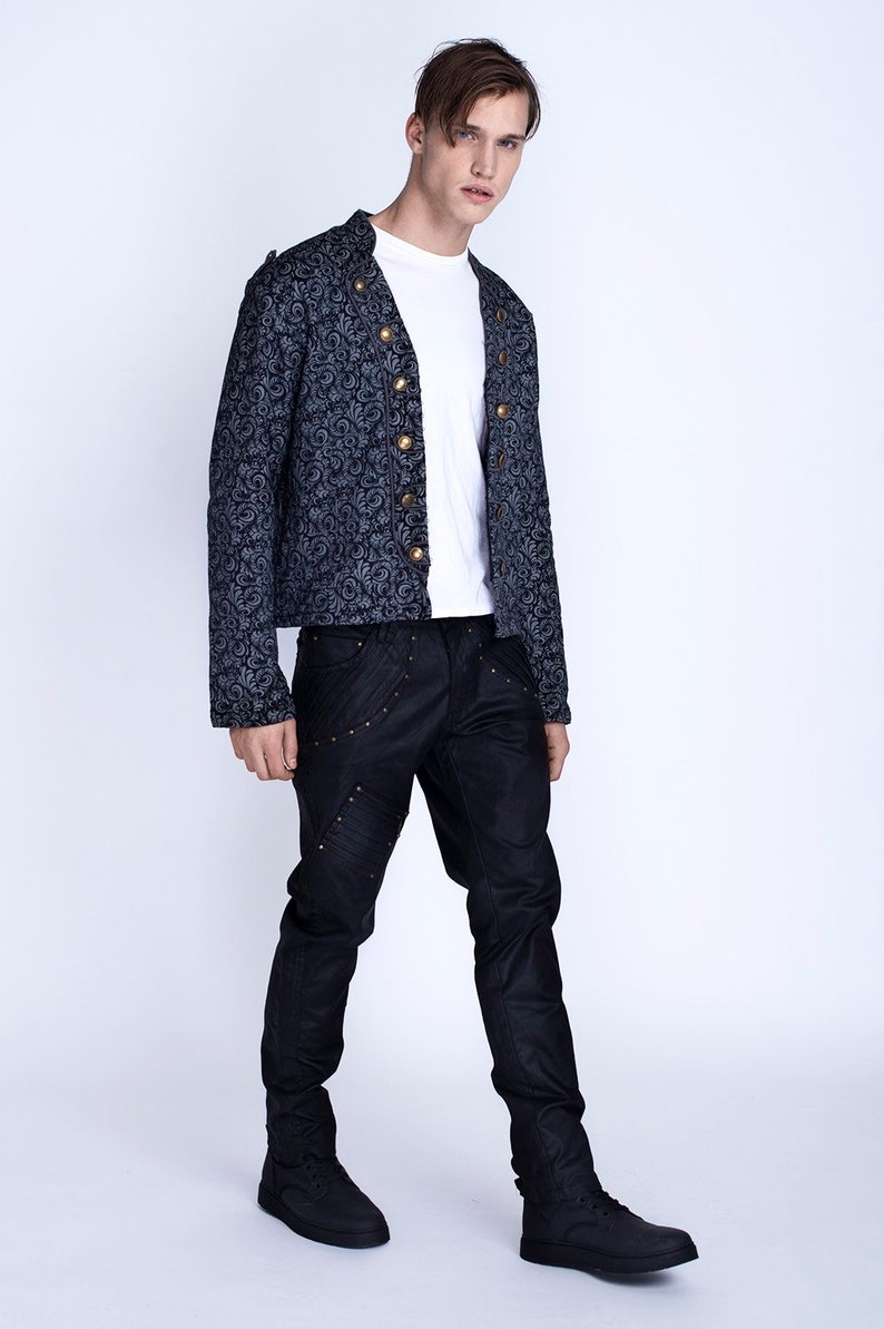 Puede incluir: Un hombre con una chaqueta estampada en negro y gris con botones dorados, pantalones vaqueros negros con costuras decorativas y zapatos negros.