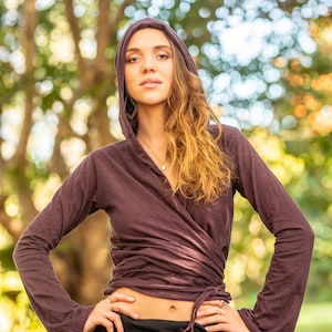 May include: A woman wearing a long-sleeved, dark purple wrap top with a hood. The top is tied at the waist and has a relaxed fit. She is wearing a black skirt with a gold geometric design.