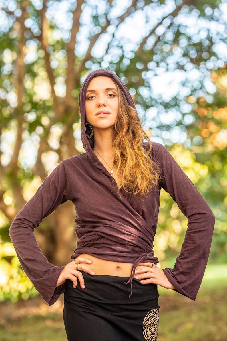 May include: A woman wearing a long-sleeved, dark purple wrap top with a hood. The top is tied at the waist and has a relaxed fit. She is wearing a black skirt with a gold geometric design.