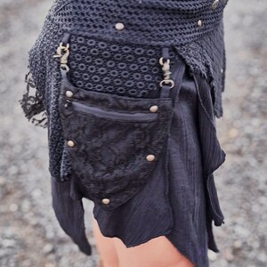 May include: Black skirt with a layered design, featuring a crochet waistband adorned with metal studs. A small, decorative pouch with a zipper and metal accents hangs from the waist. The skirt has a flowing, asymmetrical hemline.