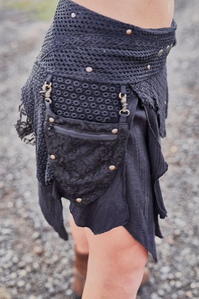 May include: Black skirt with a layered design, featuring a crochet waistband adorned with metal studs. A small, decorative pouch with a zipper and metal accents hangs from the waist. The skirt has a flowing, asymmetrical hemline.