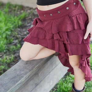 May include: A burgundy skirt with multiple layers of ruffles. The skirt has a high waist and a fitted waistband with silver buttons. The skirt is asymmetrical, with the back being longer than the front.