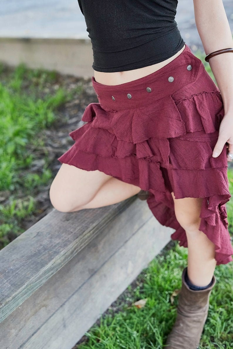 May include: A burgundy skirt with multiple layers of ruffles. The skirt has a high waist and a fitted waistband with silver buttons. The skirt is asymmetrical, with the back being longer than the front.