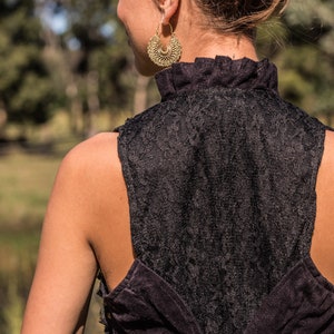 May include: A black sleeveless top featuring a high ruffled collar and a lace back panel. The garment includes a corset-style lace-up closure with black cord threaded through metal eyelets. A large, circular, textured gold-tone hoop earring.
