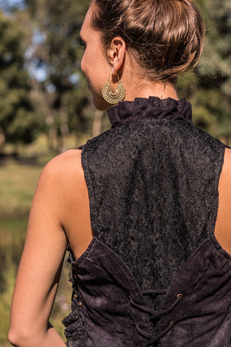 May include: A black sleeveless top featuring a high ruffled collar and a lace back panel. The garment includes a corset-style lace-up closure with black cord threaded through metal eyelets. A large, circular, textured gold-tone hoop earring.