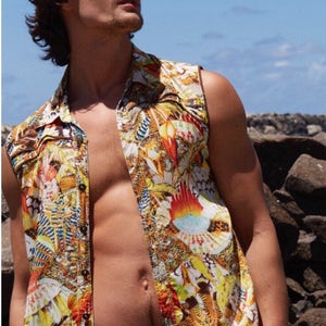May include: A sleeveless shirt with a colourful, abstract print. The shirt is unbuttoned and has a collar. The person wearing the shirt is standing in front of a rocky background.