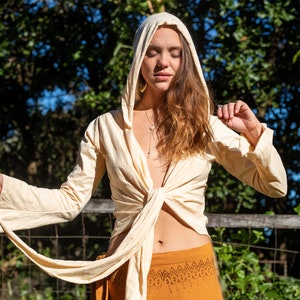 May include: A woman wearing a cream-colored hooded wrap top tied at the waist. The top has long sleeves and a relaxed fit. She is wearing a brown skirt with a geometric pattern.