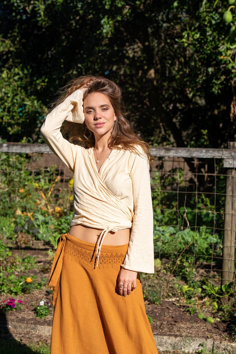 May include: A woman wearing a cream-colored wrap top with long sleeves and a mustard yellow wrap skirt with a geometric pattern. She is standing in a garden with green plants and a wooden fence in the background.