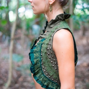 May include: A sleeveless olive green waistcoat featuring a high ruffled collar, dark green lace trim, and a vertical row of silver-toned metal studs along the side. The garment is cropped, showcasing a layered texture with fabric ruffles and lace.