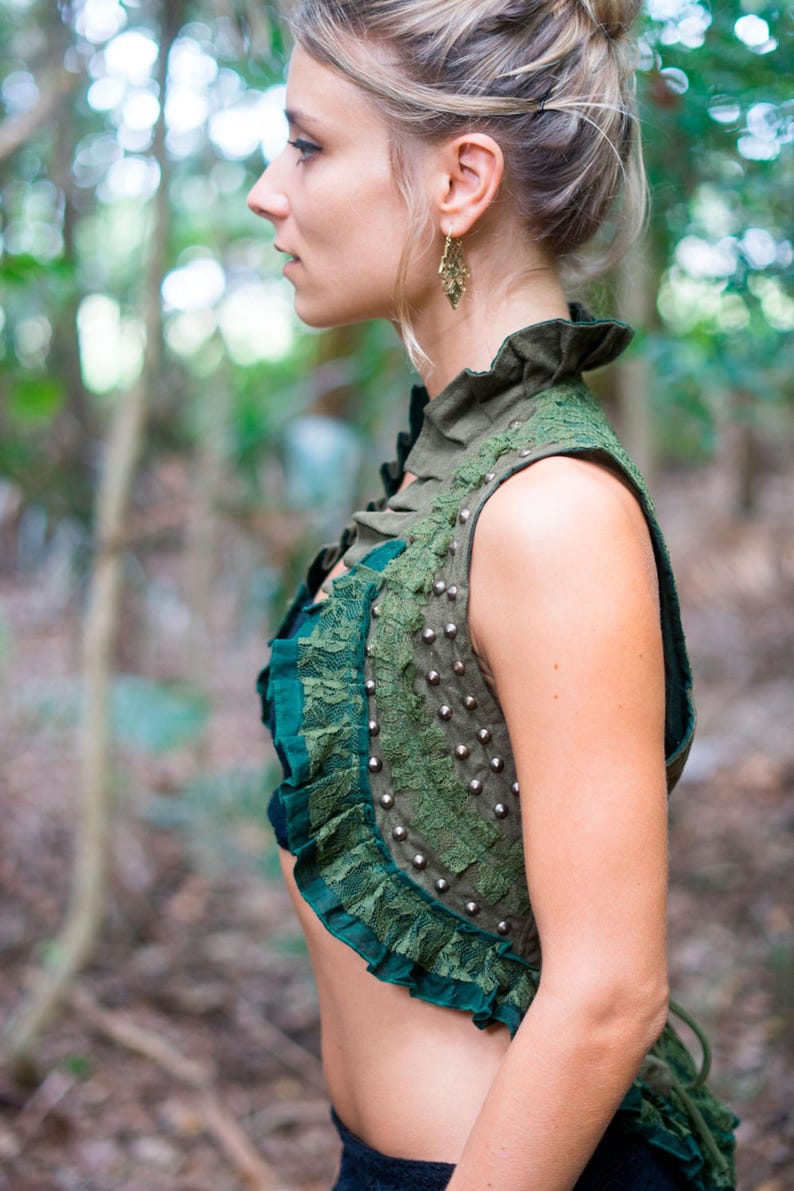 May include: A sleeveless olive green waistcoat featuring a high ruffled collar, dark green lace trim, and a vertical row of silver-toned metal studs along the side. The garment is cropped, showcasing a layered texture with fabric ruffles and lace.
