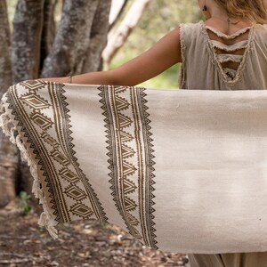 May include: A woman wearing a beige sleeveless dress with a brown and white patterned throw draped over her shoulders. The throw has a fringe border and a geometric design.