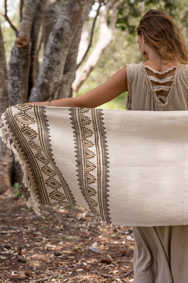 May include: A woman wearing a beige sleeveless dress with a brown and white patterned throw draped over her shoulders. The throw has a fringe border and a geometric design.