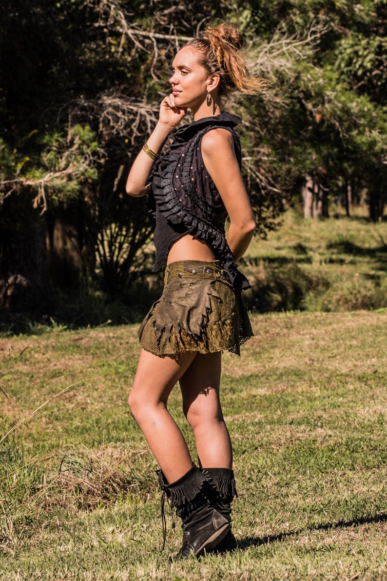 May include: A black sleeveless top with a ruffled front and a textured olive green mini skirt featuring layered fabric panels and metal studs. The outfit is paired with black mid-calf boots that have a fringed cuff detail around the top edge.