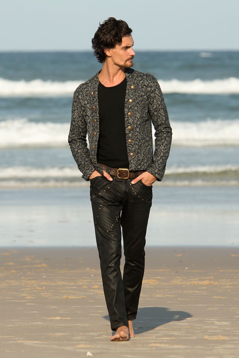 Puede incluir: Un hombre con una chaqueta negra estampada con botones dorados, pantalones vaqueros negros y un cintur&oacute;n marr&oacute;n. Est&aacute; de pie en una playa de arena con el oc&eacute;ano de fondo.