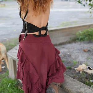 May include: A woman wearing a black halter top with a feather detail and a burgundy ruffled skirt. She is wearing brown boots and has her back to the camera.