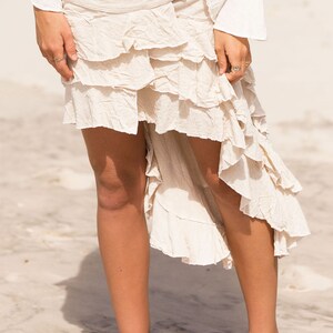 May include: A white, tiered, asymmetrical skirt with a brown fabric belt and metal accents. The skirt is made of a soft, flowing fabric and has a relaxed, bohemian style.