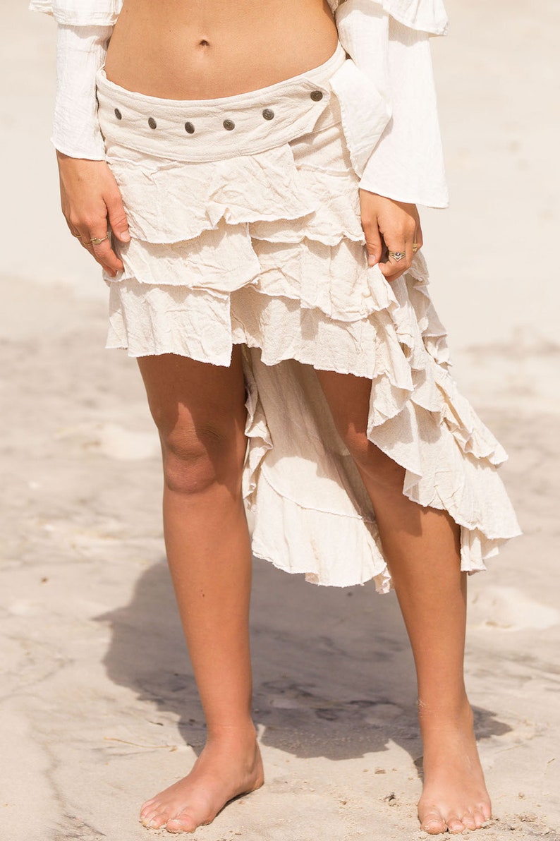 May include: A white, tiered, asymmetrical skirt with a brown fabric belt and metal accents. The skirt is made of a soft, flowing fabric and has a relaxed, bohemian style.