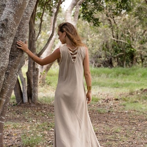 May include: A woman wearing a long, light brown linen dress with a unique criss-cross design on the back. She is standing in a forest setting, with trees and green grass in the background.