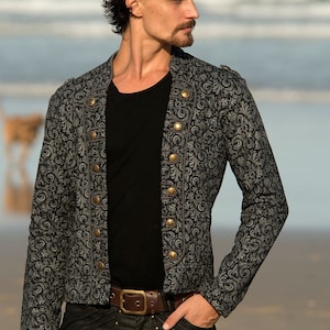 Puede incluir: Una chaqueta gris con estampado y botones dorados, con una camiseta negra debajo. La chaqueta tiene un cuello alto y mangas largas. La persona lleva pantalones negros con un cintur&oacute;n marr&oacute;n.