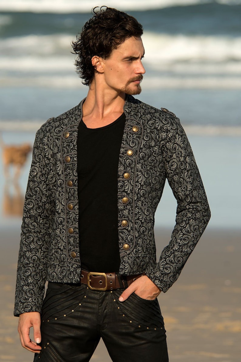 Puede incluir: Una chaqueta gris con estampado y botones dorados, con una camiseta negra debajo. La chaqueta tiene un cuello alto y mangas largas. La persona lleva pantalones negros con un cintur&oacute;n marr&oacute;n.