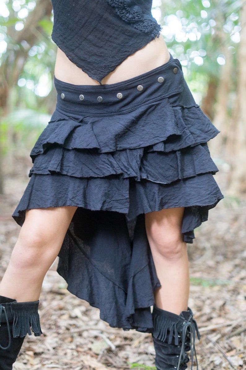 May include: A black, tiered skirt with a high-low hemline. The skirt has a wide waistband with silver buttons and a ruffled bottom.