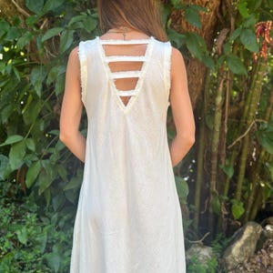 May include: A white sleeveless maxi dress with a V-neckline and a unique criss-cross strap detail on the back. The straps are frayed and create an open back design.