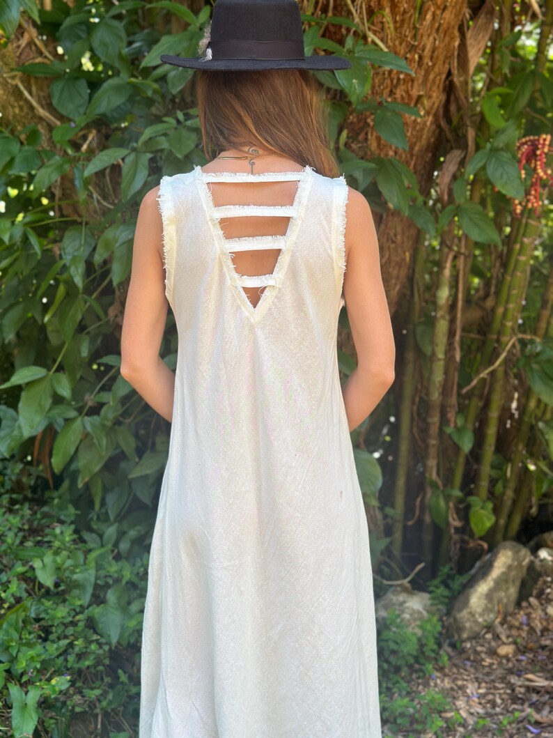 May include: A white sleeveless maxi dress with a V-neckline and a unique criss-cross strap detail on the back. The straps are frayed and create an open back design.