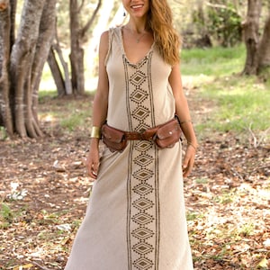 May include: A woman wearing a beige maxi dress with a brown geometric pattern. She is wearing a brown leather belt with multiple pockets and a gold bracelet on her left wrist. She is standing in a forest setting.