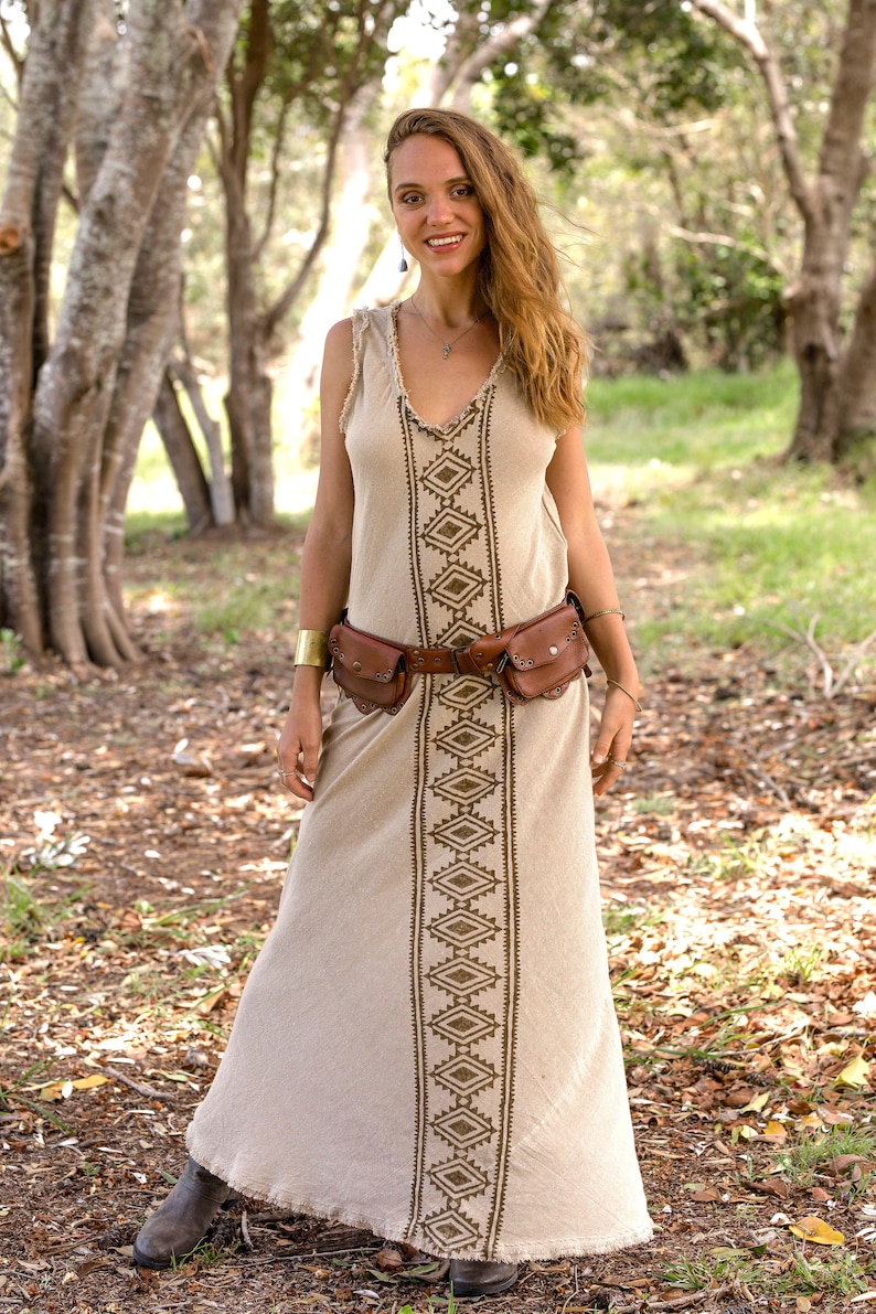 May include: A woman wearing a beige maxi dress with a brown geometric pattern. She is wearing a brown leather belt with multiple pockets and a gold bracelet on her left wrist. She is standing in a forest setting.