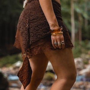 May include: A brown lace skirt with a layered design. The skirt has an asymmetrical hemline and is paired with a matching brown top. The outfit is accessorized with amber bracelets and rings.
