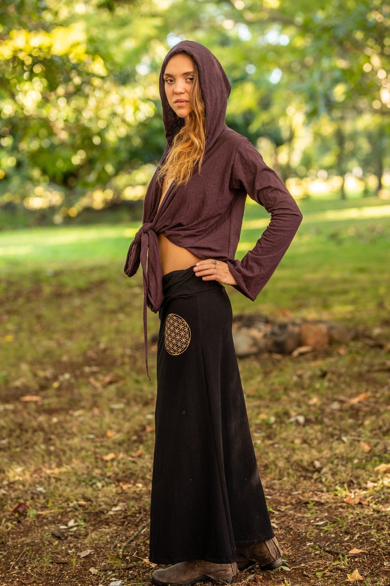 May include: A woman wearing a long, black skirt with a gold flower of life design. She is also wearing a purple hooded top tied at the waist.