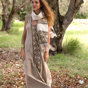 May include: A woman wearing a long, beige linen dress with brown geometric embroidery. She is wearing a white and brown patterned scarf with fringe and a gold bracelet. The dress is flowing in the wind and she is standing in a forest setting.