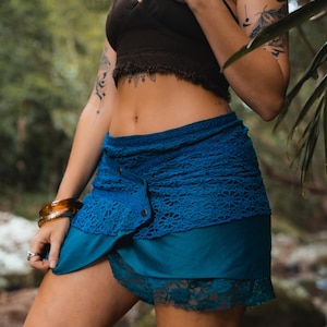 May include: A teal and blue wrap skirt with a lace overlay and button closure. The skirt is paired with a dark brown halter top. The image is taken outdoors with a blurred green background.