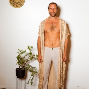 Puede incluir: Un hombre con una t&uacute;nica beige con estampado y ribete marr&oacute;n sobre un pantal&oacute;n beige claro. Est&aacute; de pie sobre una alfombra estampada frente a una pared blanca.