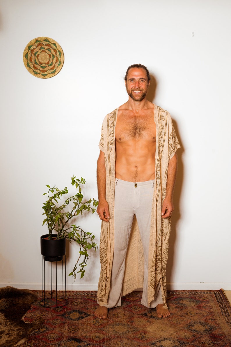 Puede incluir: Un hombre con una t&uacute;nica beige con estampado y ribete marr&oacute;n sobre un pantal&oacute;n beige claro. Est&aacute; de pie sobre una alfombra estampada frente a una pared blanca.