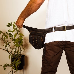 K&ouml;nnte beinhalten: Eine schwarze Leder-Bauchtasche mit einem Rei&szlig;verschlussfach und Metallnieten. Die Bauchtasche wird um die Taille einer Person getragen, die eine braune Shorts und ein wei&szlig;es T-Shirt tr&auml;gt.